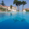 Piscina all'aperto con vista su edifici bianchi e alberi alti in lontananza, riflessi nell'acqua.